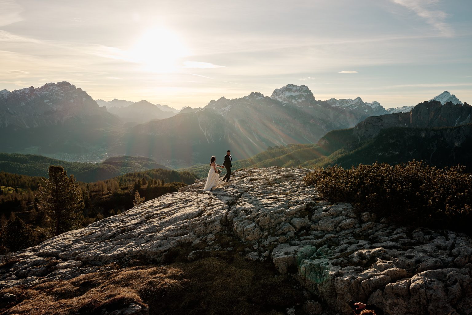 Elisa and Hector small against a wide Dolomites sunrise, sunburst over the ridge
