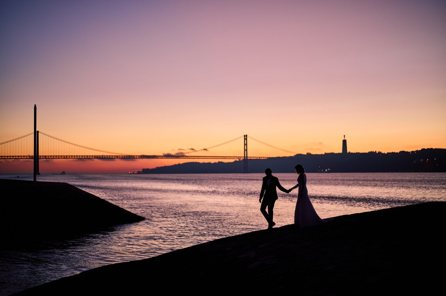 Ana and David walking hand in hand on the breakwater beneath the 25 de Abril bridge at dawn
