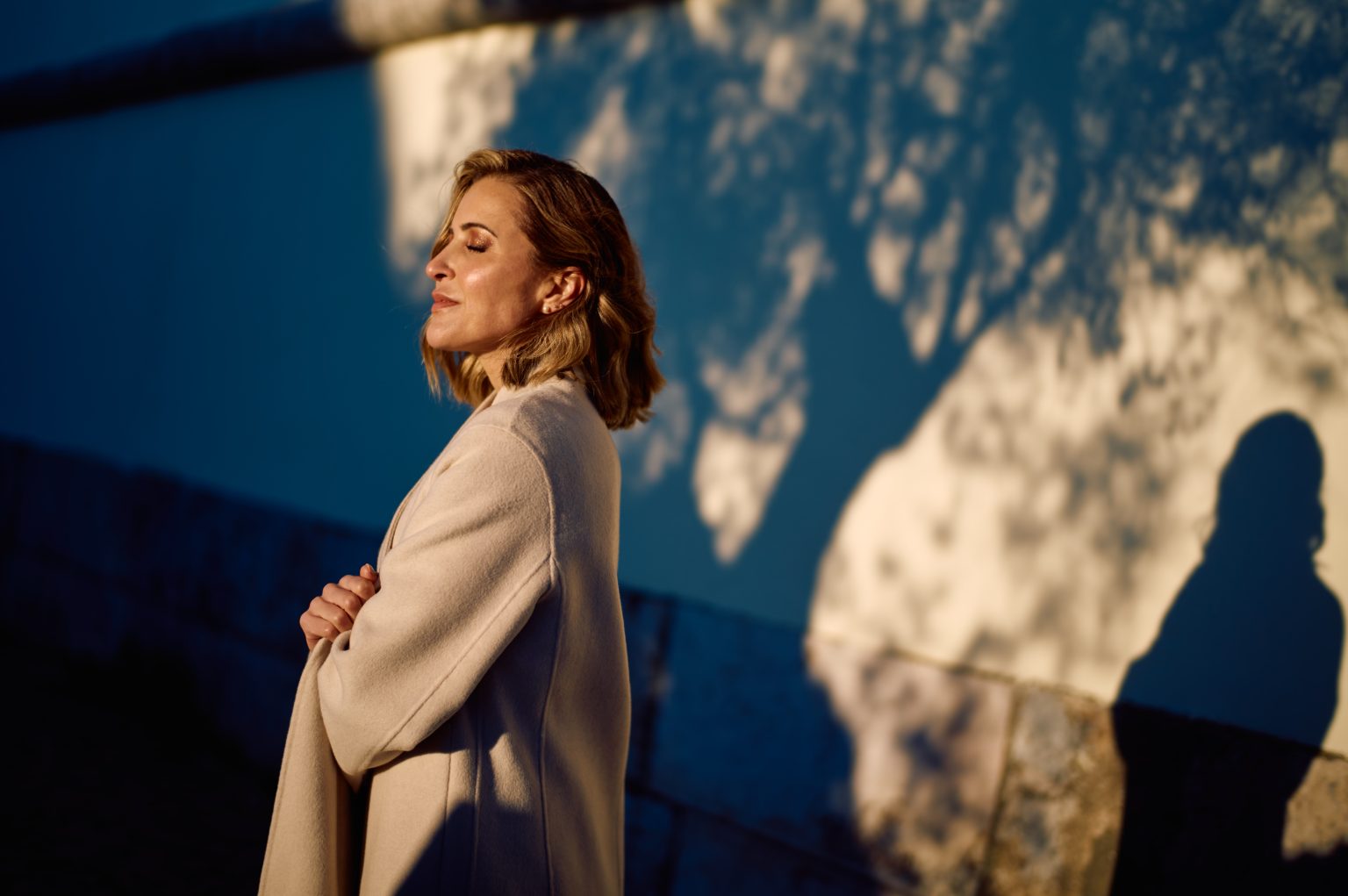 Ana in her coat against a blue wall with morning tree shadows in Lisbon