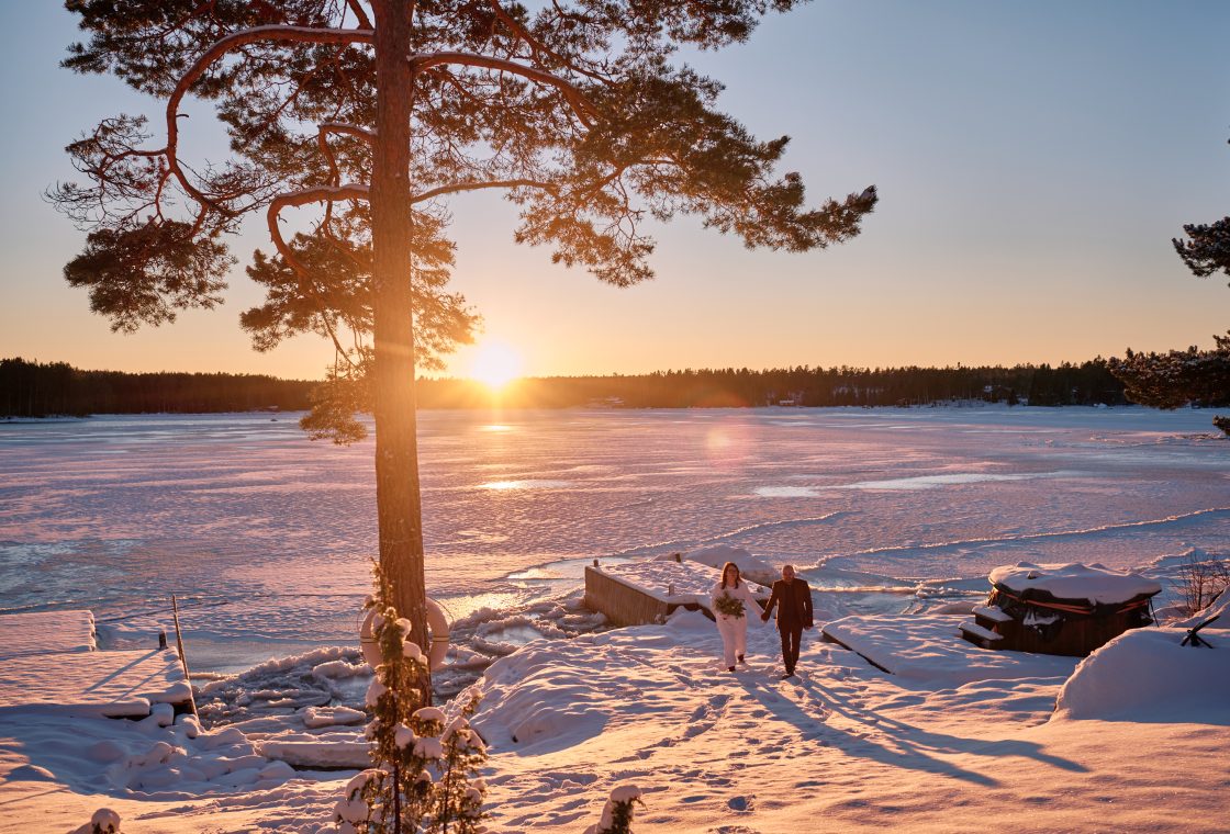 Sam and Rob walking the frozen shore at sunset by the cabin, a winter elopement in Sweden