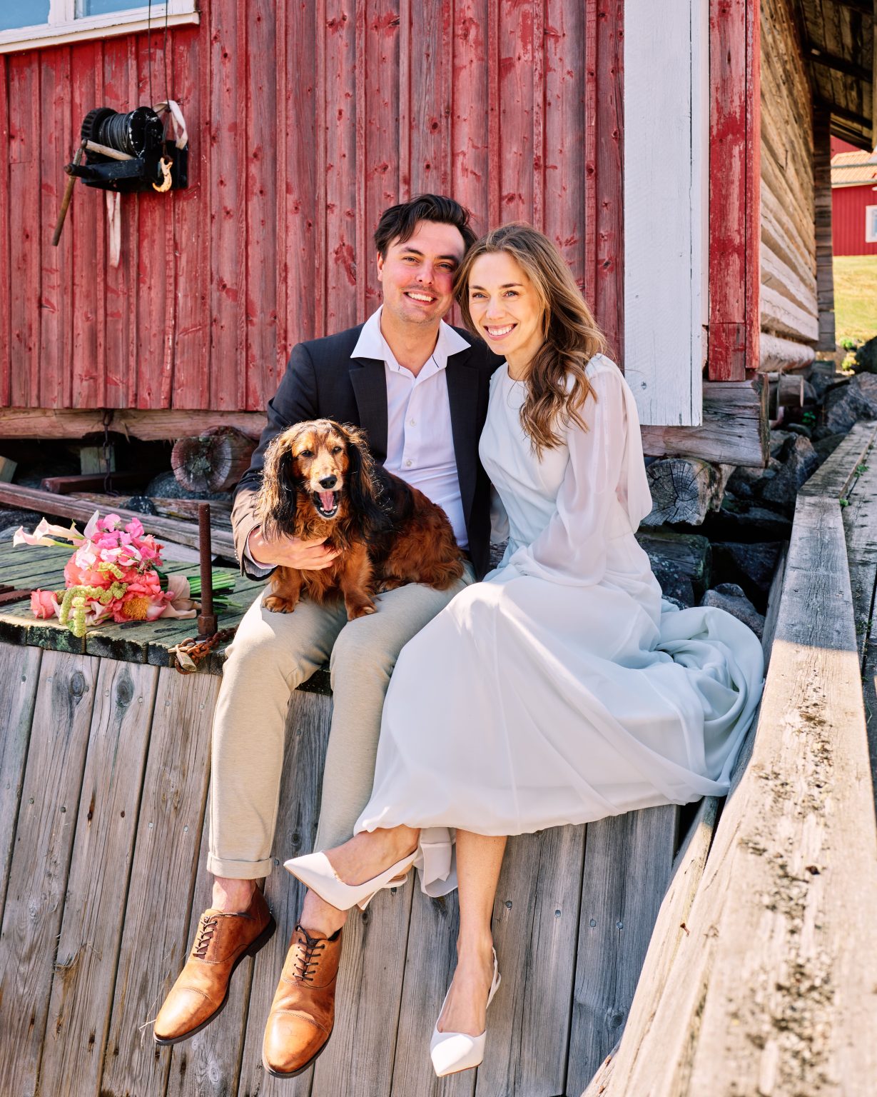 Elin and Joakim with their dog on a wooden deck by a red barn, Skärså