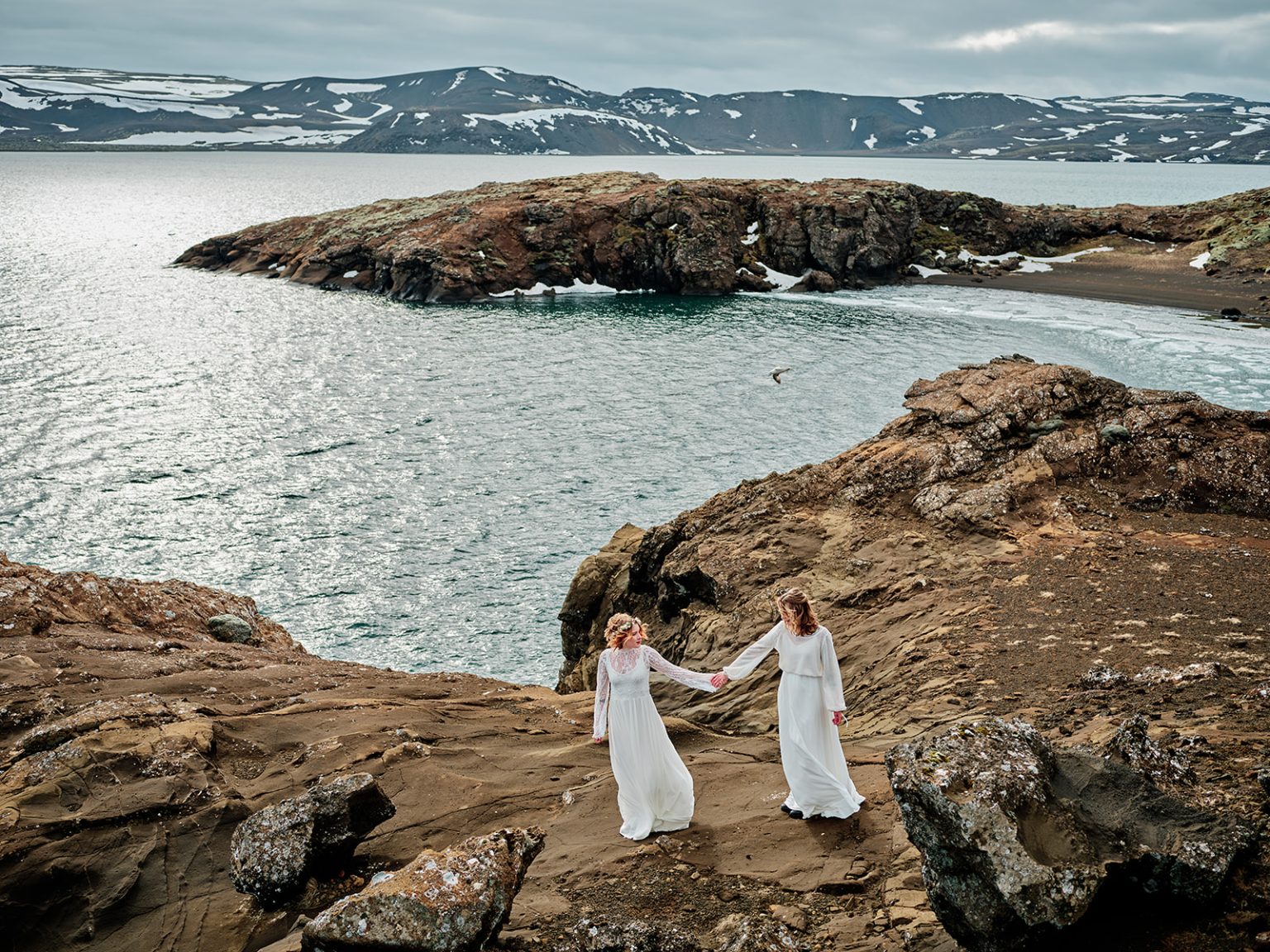 Two brides hand in hand above a blue crater lake, gay elopement in Iceland