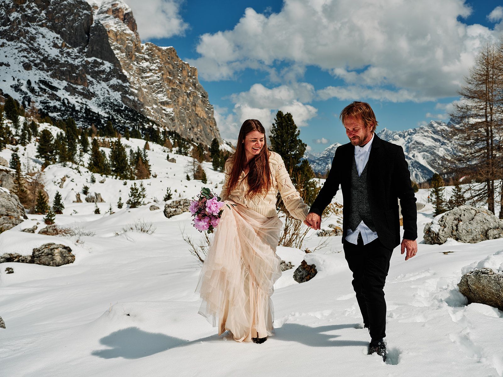 Bride and groom walking with the bouquet past a Dolomites chapel at Passo Gardena - Nordica Photography